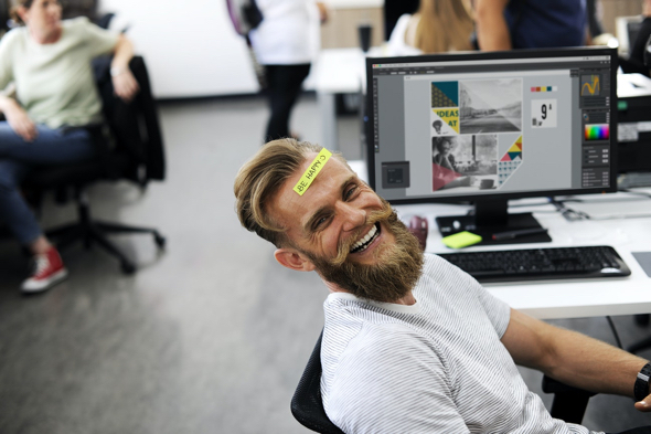 laughing guy with blond hair beard at computer with post-it note on his forehead which says Be Happy