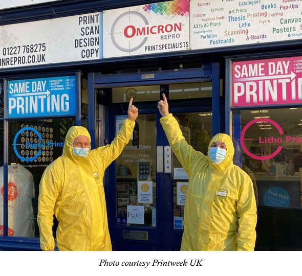 Owner Mark Fawcett-Jones and and director Dave Loveridge wearing hazmat suits and pointing to their store sign, Omicron Repro, Print Specialists in Canterbury, England