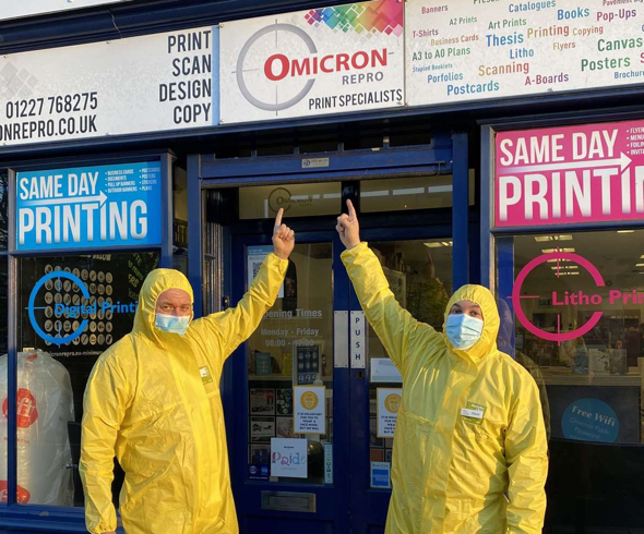 Owner Mark Fawcett-Jones and and director Dave Loveridge wearing hazmat suits and pointing to their store sign, Omicron Repro, Print Specialists in Canterbury, England